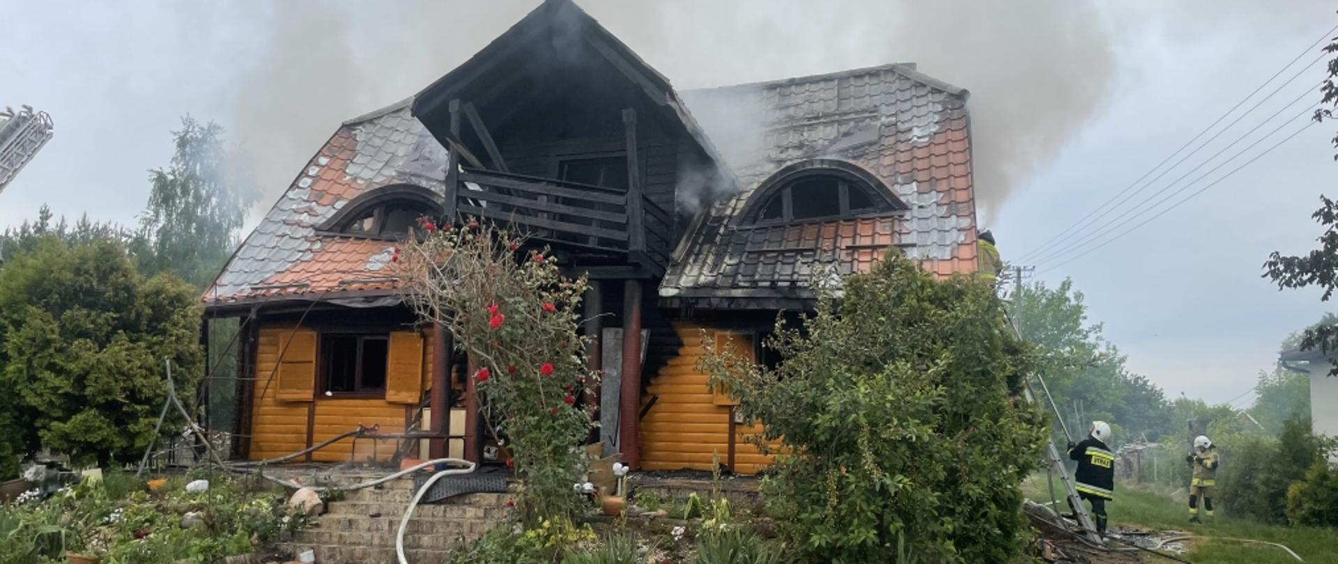 Widok od frontu na spalony budynek mieszkalny konstrukcji drewnianej. Po prawej stronie kadru widoczni dwaj strażacy OSP. Przed budynkiem porozwijane linie wężowe napełnione wodą. 