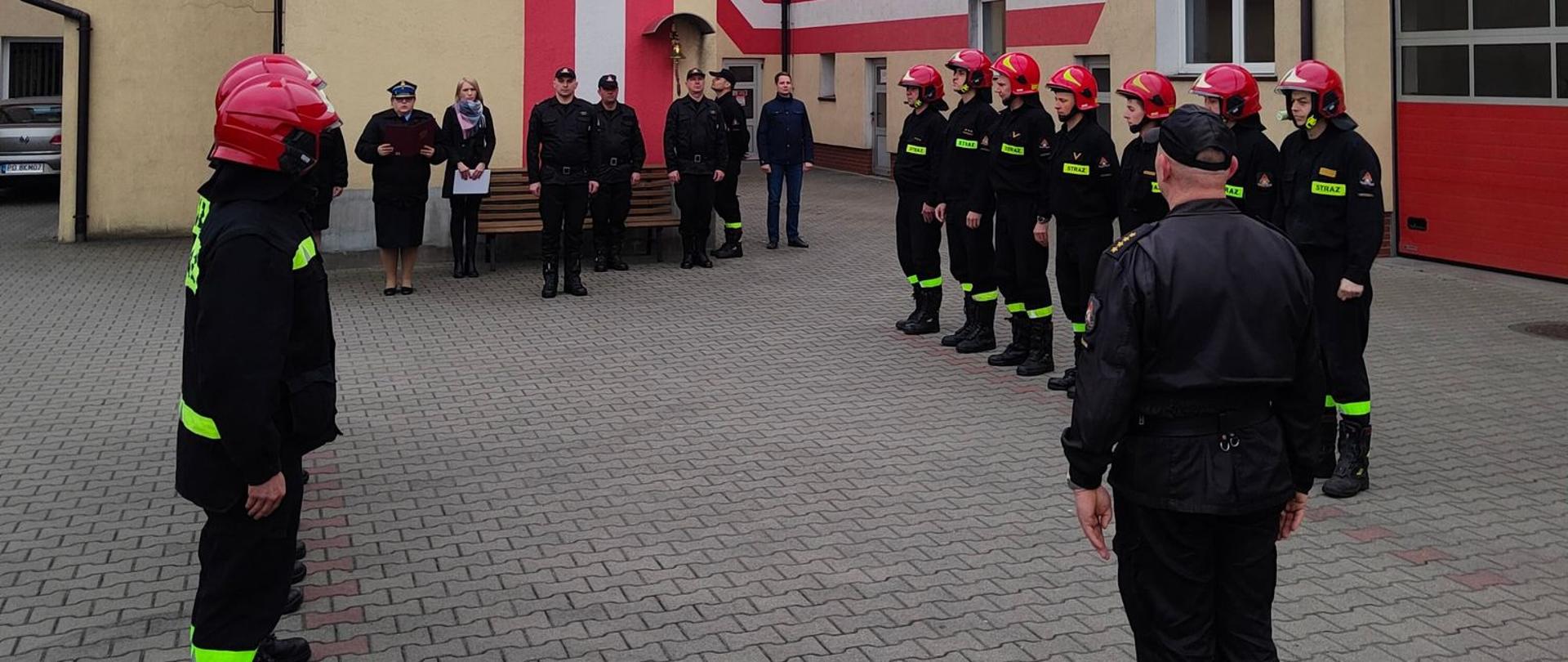 Zdjęcie przedstawia Strażaków Komendy Powiatowej PSP w Krotoszynie podczas zmiany służby. Strażacy stoją w dwóch szeregach po przeciwległej stronie. Prostopadle do strażaków pełniących służbę w podziale bojowym stoją funkcjonariusze systemu codziennego i pracownicy cywilni Komendy. 