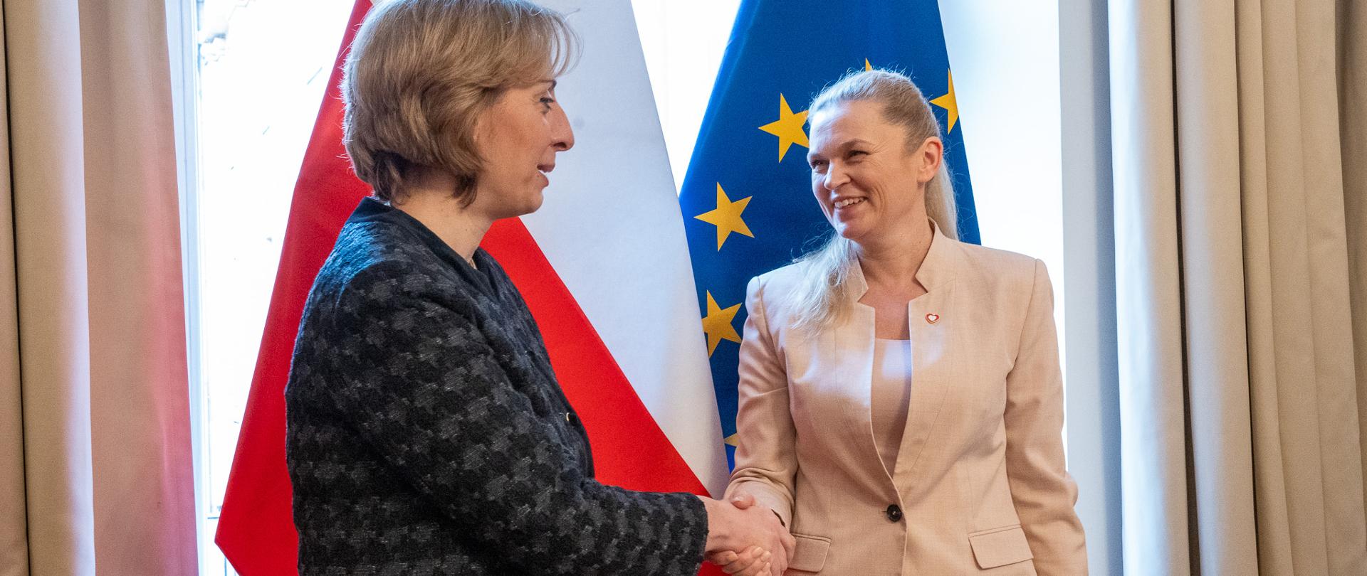 Minister Barbara Nowacka podaje rękę kobiecie w ciemnej garsonce. W tle flagi Polski i UE
