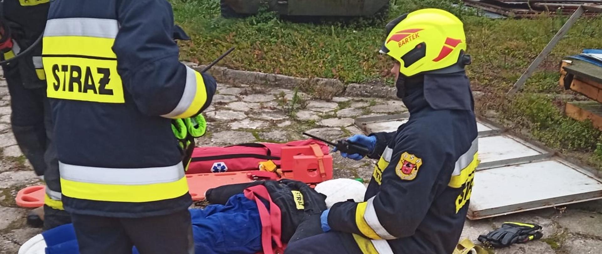Zdjęcie przedstawia strażaków podczas ćwiczeń, zainscenizowane stanowisko pomocy medycznej, na noszach widać pozoranta, wokół rozłożone torby medyczne, na pierwszym planie 2 strażaków w umundurowaniu i żółtych kaskach