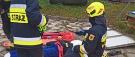 Zdjęcie przedstawia strażaków podczas ćwiczeń, zainscenizowane stanowisko pomocy medycznej, na noszach widać pozoranta, wokół rozłożone torby medyczne, na pierwszym planie 2 strażaków w umundurowaniu i żółtych kaskach