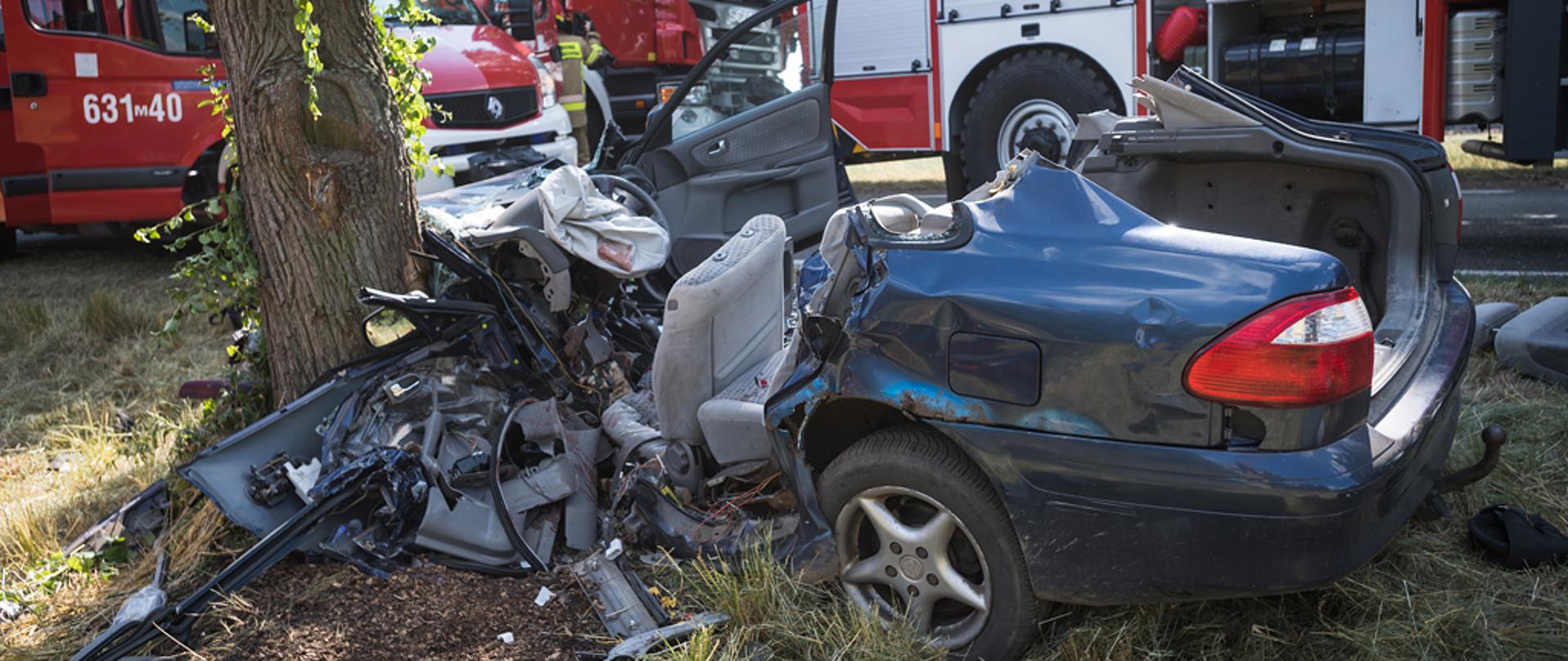 Na zdjęciu widoczny samochód osobowy ciemnego koloru roztrzaskany na przydrożnym drzewie. Auto w wyniku działań strażaków pozbawione dachu. Na drugim planie stoją samochody straży pożarnej.