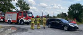 Na zdjęciu znajdują się stojący strażacy Państwowej Straży Pożarnej. Z lewej strony zaparkowane są 2 wozy strażackie. Z prawej strony stoi czarny OPEL po uprzednim zderzeniu. 