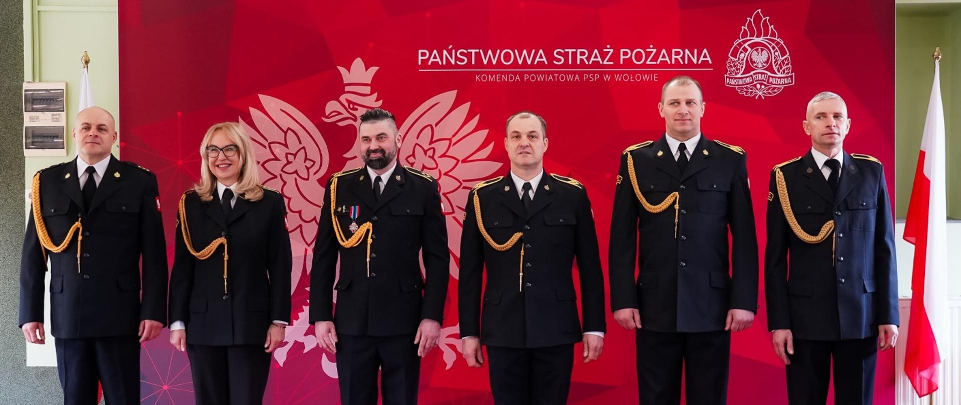 Zdjęcie przedstawia sześć osób w oficjalnych mundurach strażackich, stojących w rzędzie na tle czerwonej ścianki reklamowej W rzędzie stoi pięciu mężczyzn i jedna kobieta. Wszyscy są uśmiechnięci lub zachowują powagę godną uroczystości. Każda z osób ma na sobie czarny mundur wyjściowy Państwowej Straży Pożarnej. Mundury ozdobione są złotymi sznurami galowymi przypiętymi do prawego ramienia. Na ramionach widoczne są pagony z oznaczeniami stopni służbowych. Ścianka: Ma intensywny czerwony kolor z grafiką przedstawiającą białego orła w koronie (godło Polski) oraz napisy. Główny napis głosi: „Państwowa Straż Pożarna”, a poniżej mniejszym drukiem: „Komenda Powiatowa PSP W Wołowie”. Osoby stoją na czerwonym dywanie, który kolorystycznie współgra z tłem. Po prawej stronie kadru widoczna jest flaga Polski na metalowym stojaku.