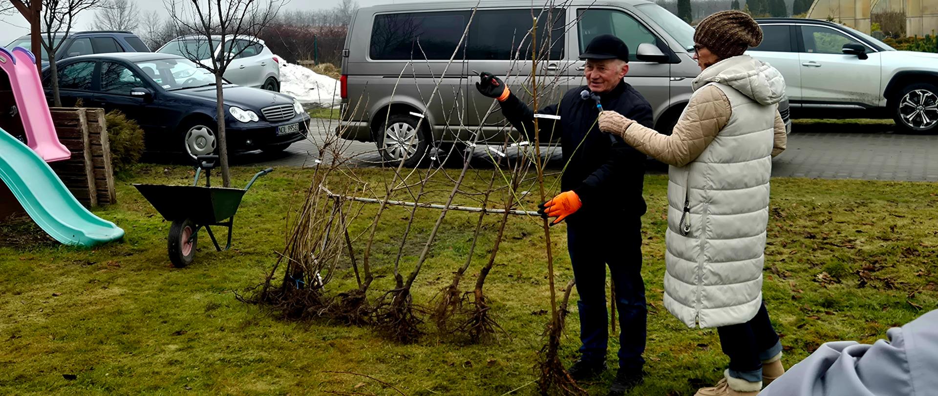 Mężczyzna w czarnej kurtce i pomarańczowymi rękawicami mówi do mikrofonu, który trzyma kobieta w białej kurtce. W tle zaparkowane samochody