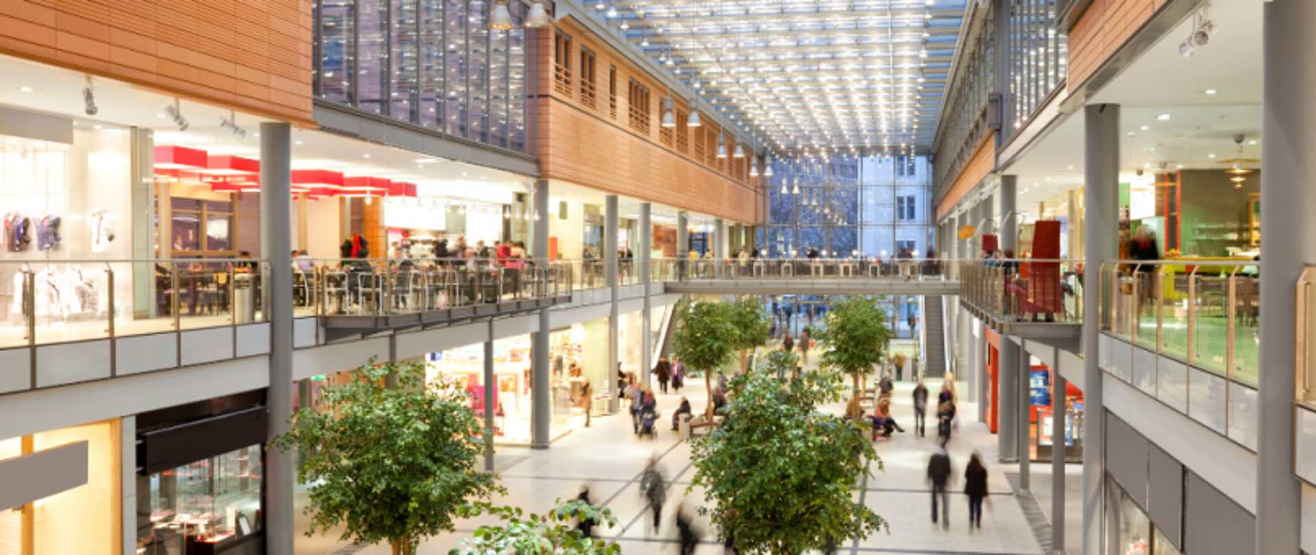 Dwupoziomowe centrum handlowe z szklanym dachem, sklepami, strefą gastronomiczną i roślinnością w donicach.