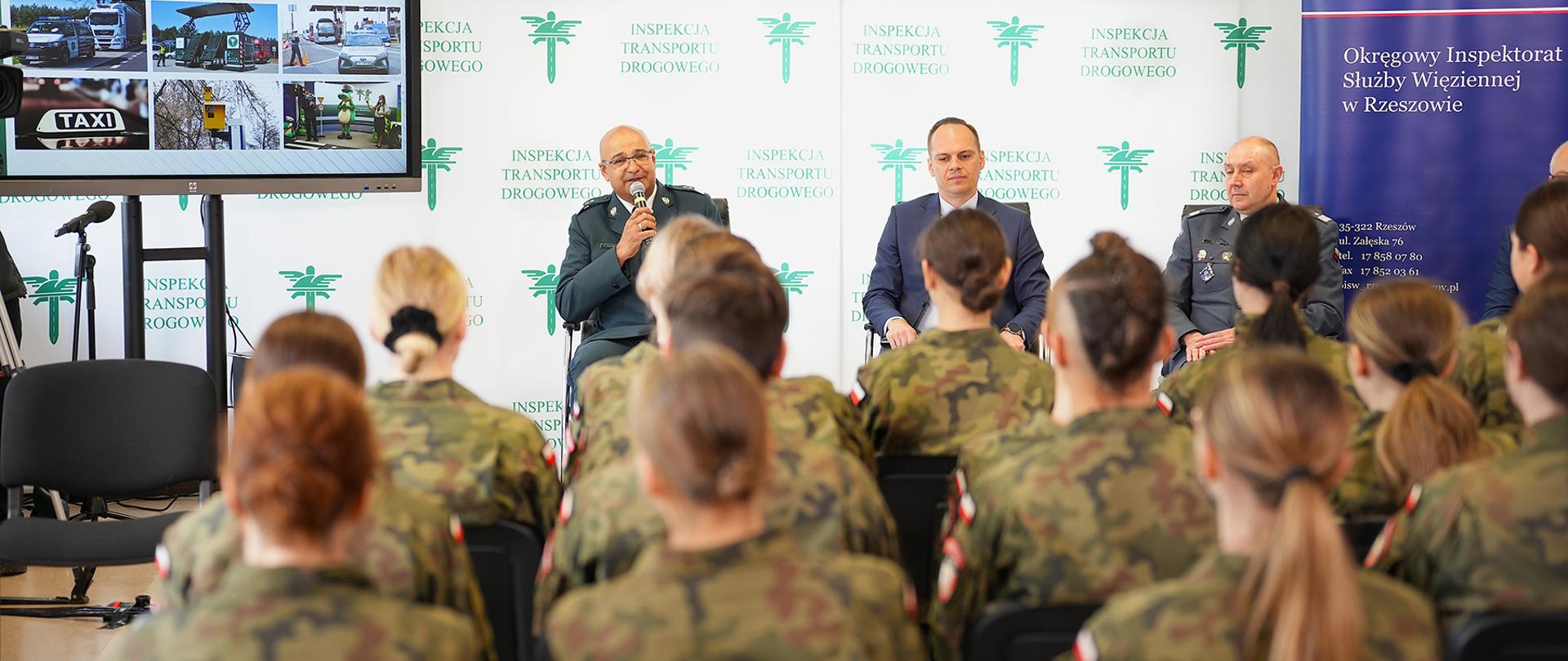 Szef ITD zaznaczył, że wskaźnik wypadkowości wśród młodych kierowców (w grupie wiekowej 18-24) jest najwyższy, jeśli chodzi o całą populację. Lekcje brd w ramach projektu edukacyjnego „Szanuj zasady, chroń życie” służą temu, by młodych kierowców przestrzegać przed brawurą. Trwa lekcja. Na pierwszym planie odwróceni plecami uczniowie, słuchają. W centrum zdjęcia Szef ITD, Wiceminister MI i Dyrektor Generalny SW, mówi Szef ITD, trzyma mikrofon. W tle ścianka ITD i SW. Po lewej stronie zdjęcia telewizor z prezentacją. Pomieszczenie to sala w zakładzie karnym.