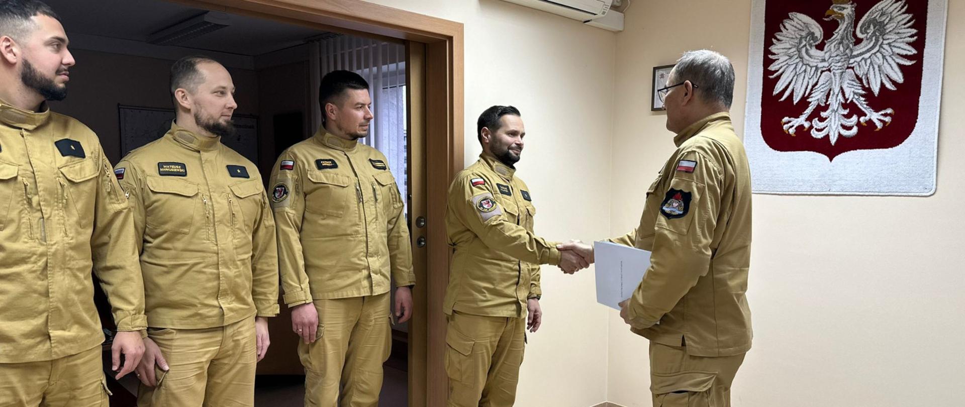 W jasnym pomieszczeniu czterech mężczyzn w beżowych mundurach stoi w szeregu przy wejściu do pokoju. Jeden z nich, stojący najbliżej prawej strony, ściska dłoń mężczyzny zwróconego do kamery plecami, który również ma mundur i trzyma teczkę z dokumentami. Na ścianie po prawej widnieje duży wizerunek polskiego orła państwowego.