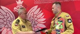 Komendant Powiatowy PSP wręcza wyróżnienie funkcjonariuszowi. Strażacy w piaskowych mundurach podają sobie dłonie na tle ścianki Państwowej Straży Pożarnej.