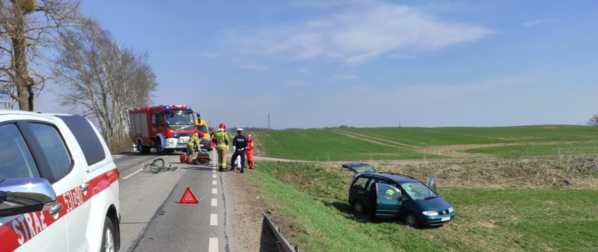 Służby ratunkowe udzielają pomocy rannemu mężczyźnie, który leży na poboczu drogi. Na drodze stoją samochody Państwowej Straży Pożarnej, a poza drogą stoi samochód osobowy, który uderzył w rowerzystę. 