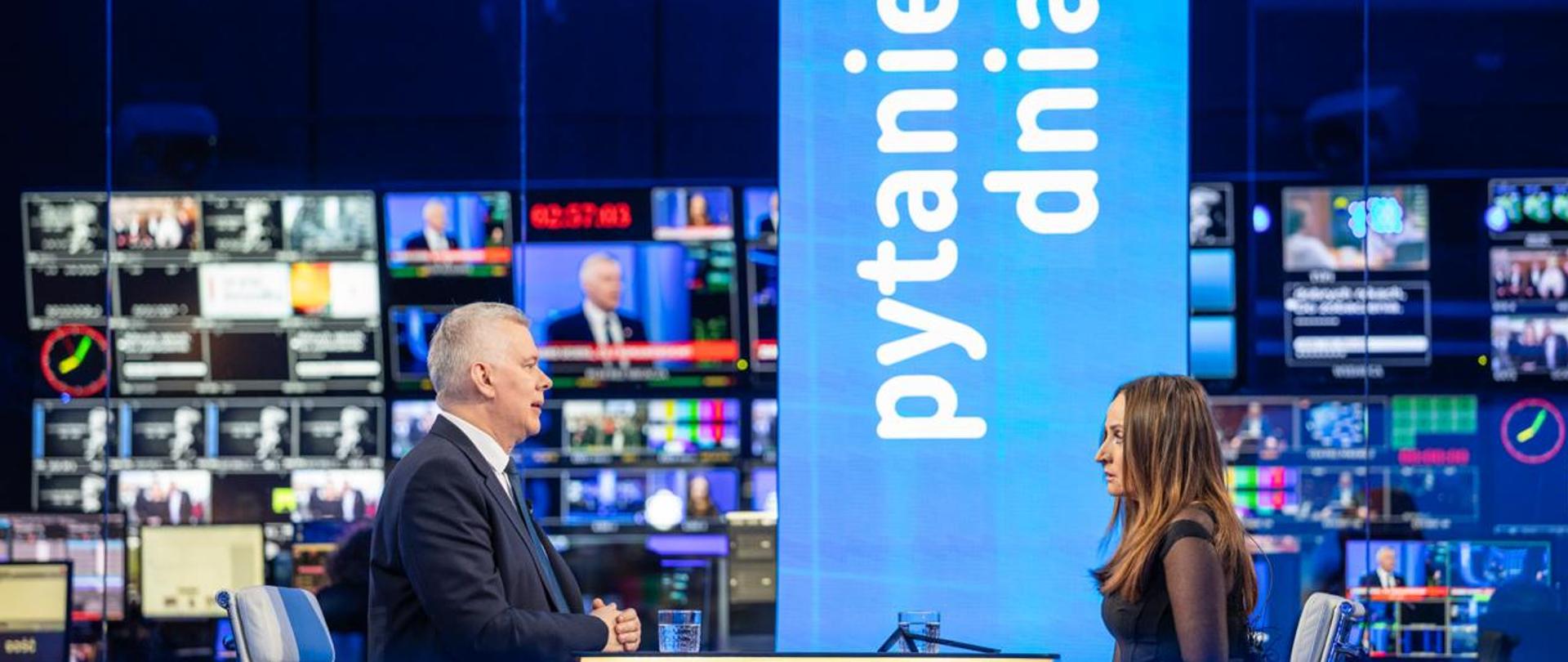 Na zdjęciu wnętrze studia telewizyjnego TVP INFO. Przy stole na białych krzesłach siedzą: minister Tomasz Siemoniak (po lewej) oraz redaktor Justyna Dobrosz-Oracz (po prawej). Na stole szklanki z wodą. Za stołem widać poziomy wyświetlacz z białym napisem "pytanie dnia" na niebieskim tle plus czerwony prostokąt. W tle widać wiele telewizyjnych monitorów.