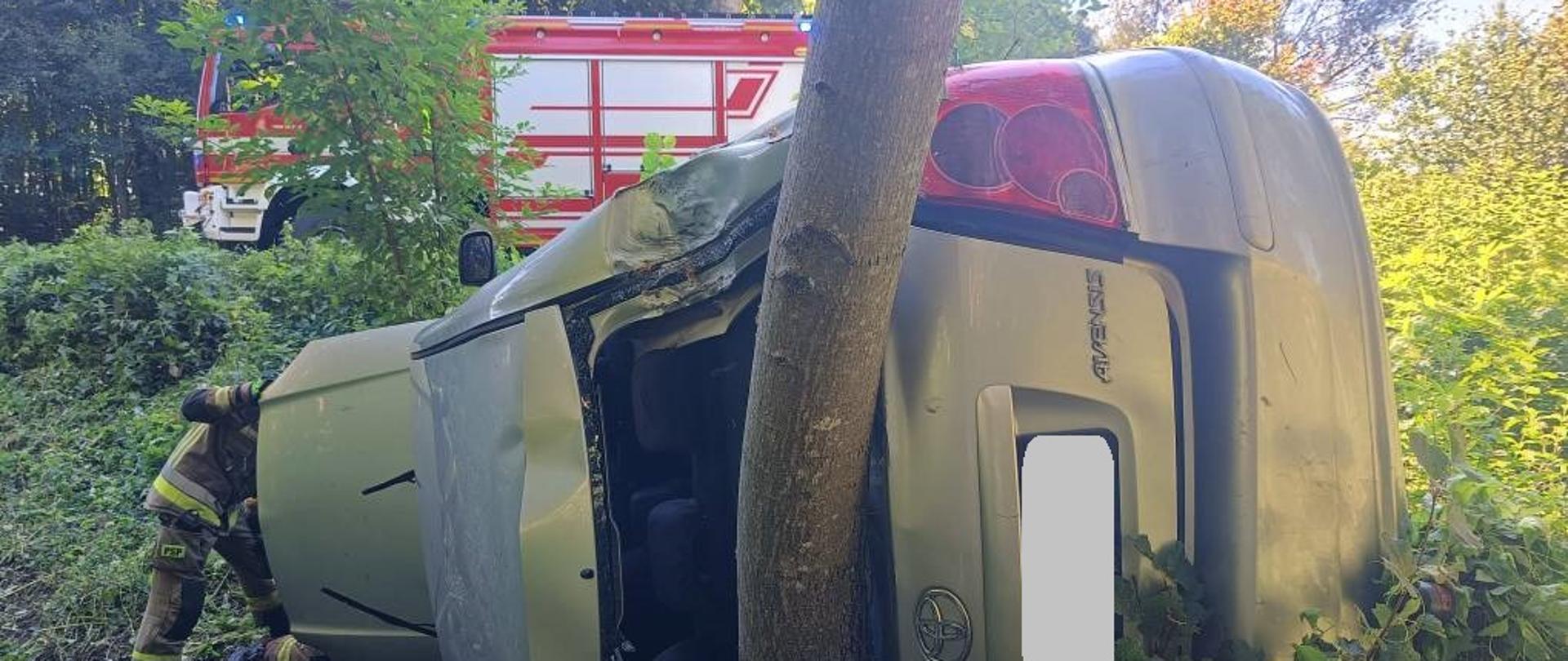 Jasny samochód osobowy leży na boku poza droga po uderzeniu w drzewo. U góry stoi samochód strażacki.