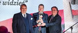 Minister Czarnek stoi między dwoma mężczyznami w garniturach, wszyscy razem uśmiechają się i trzymają w rękach statuetkę, za nimi biało-czerwona ściana.