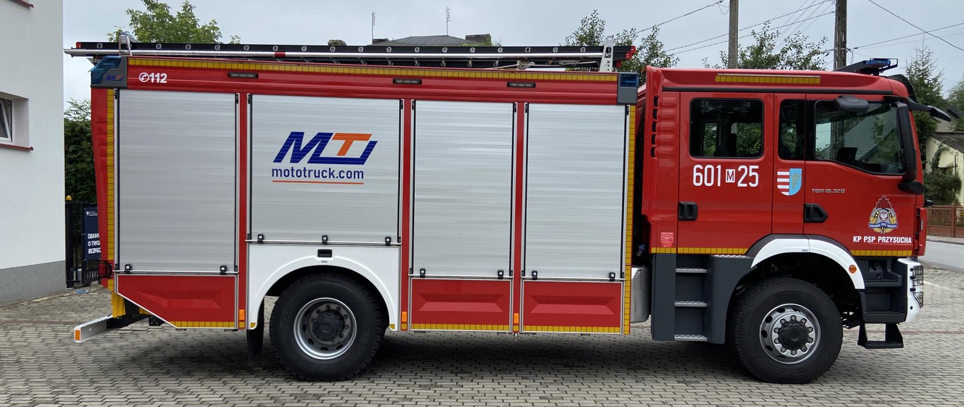 Zdjęcie przedstawia duży, nowoczesny wóz strażacki koloru czerwonego z białymi i srebrnymi elementami, stojący na wybrukowanym placu przed budynkiem w Przysusze. Na boku pojazdu widoczny jest numer "601-25", logo "MTI mototruck.com" oraz napis "KP PSP PRZYSUCHA", co wskazuje na to, że jest to pojazd Komendy Powiatowej Państwowej Straży Pożarnej w Przysusze. Na górze wozu znajduje się drabina. W tle widać fragmenty budynków i zieleni. Niebo jest pochmurne.