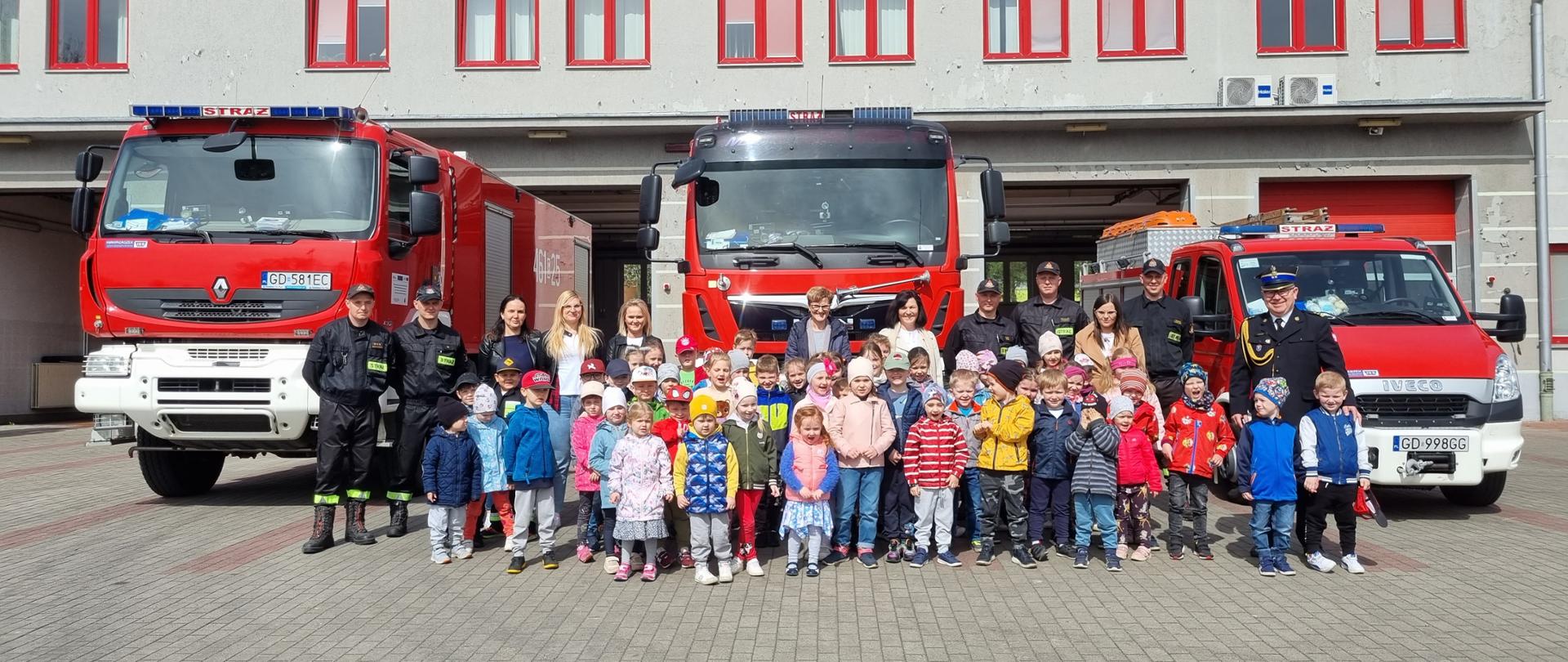 Dzieci, opiekunki oraz strażacy stoją na tle samochodów strażackich na placu KP PSP Kwidzyn
