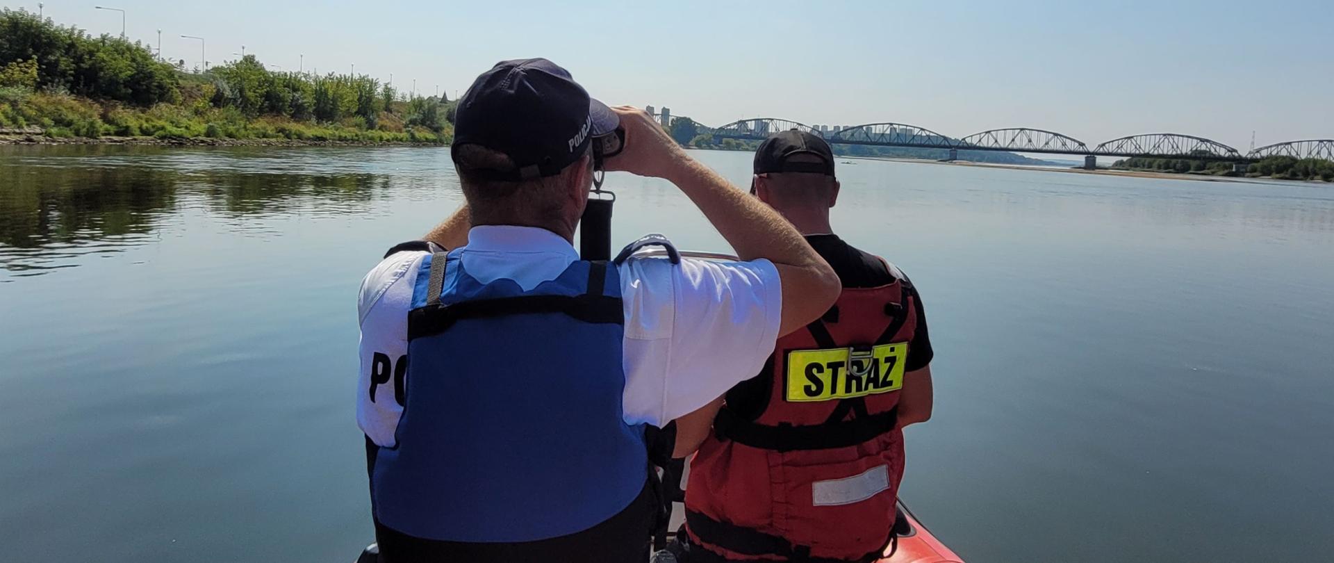 Zdjęcie przedstawia dwóch mężczyzn widzianych od tyłu na pokładzie łodzi. Obaj mają założone kamizelki asekuracyjne. Ten w niebieskiej patrzy przed siebie przez lornetkę. Dookoła nich woda. W dali widoczne zielone brzegi i most nad rzeką.