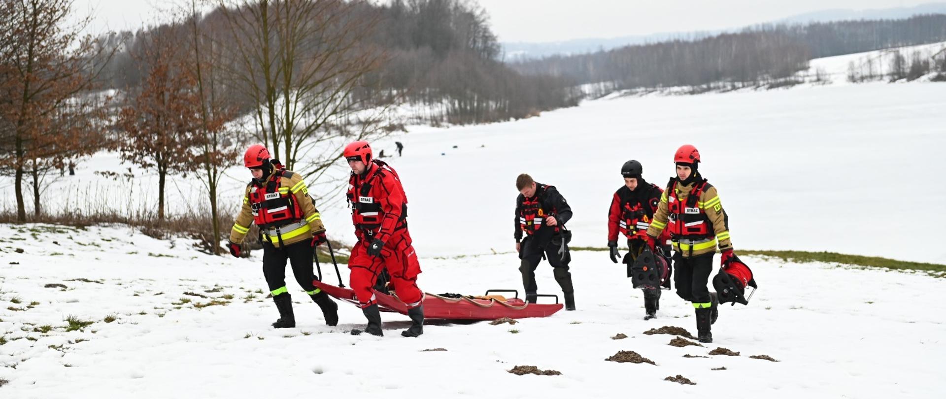 Strażacy ciągną po śniegu czerwone sanie lodowe. Mają na sobie kamizelki wypornościowe, a głowach mają kaski. W tle drzewa oraz jezioro pokryte lodem i śniegiem