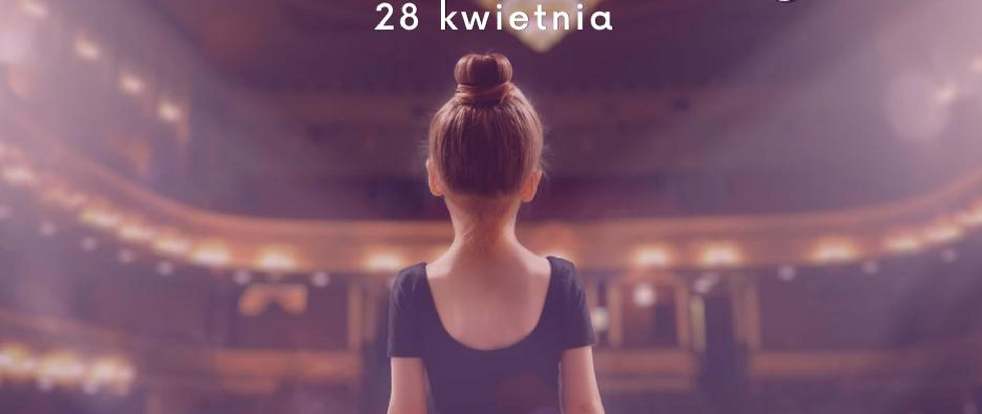 Dziewczynka w czarnej koszulce i różowej spódniczce baletowej stoi odwrócona tyłem. Ma ciemne włosy upięte w koczek. W tle sala baletowa lub operowa z świecącymi reflektorami.