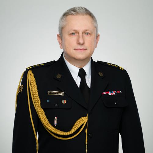 Mężczyzna w średnim wieku ubrany w galowy mundur Państwowej Straży Pożarnej stoi na neutralnym, jasnym tle. Ma krótkie, siwe włosy i poważny, spokojny wyraz twarzy. Na ciemnym mundurze widoczne są oznaczenia stopnia na pagonach, baretki odznaczeń, przypinki oraz imiennik z napisem S. Sierpatowski. Przez ramię przewieszony jest ozdobny, złoty sznur galowy. Zdjęcie ma charakter oficjalnego portretu służbowego.