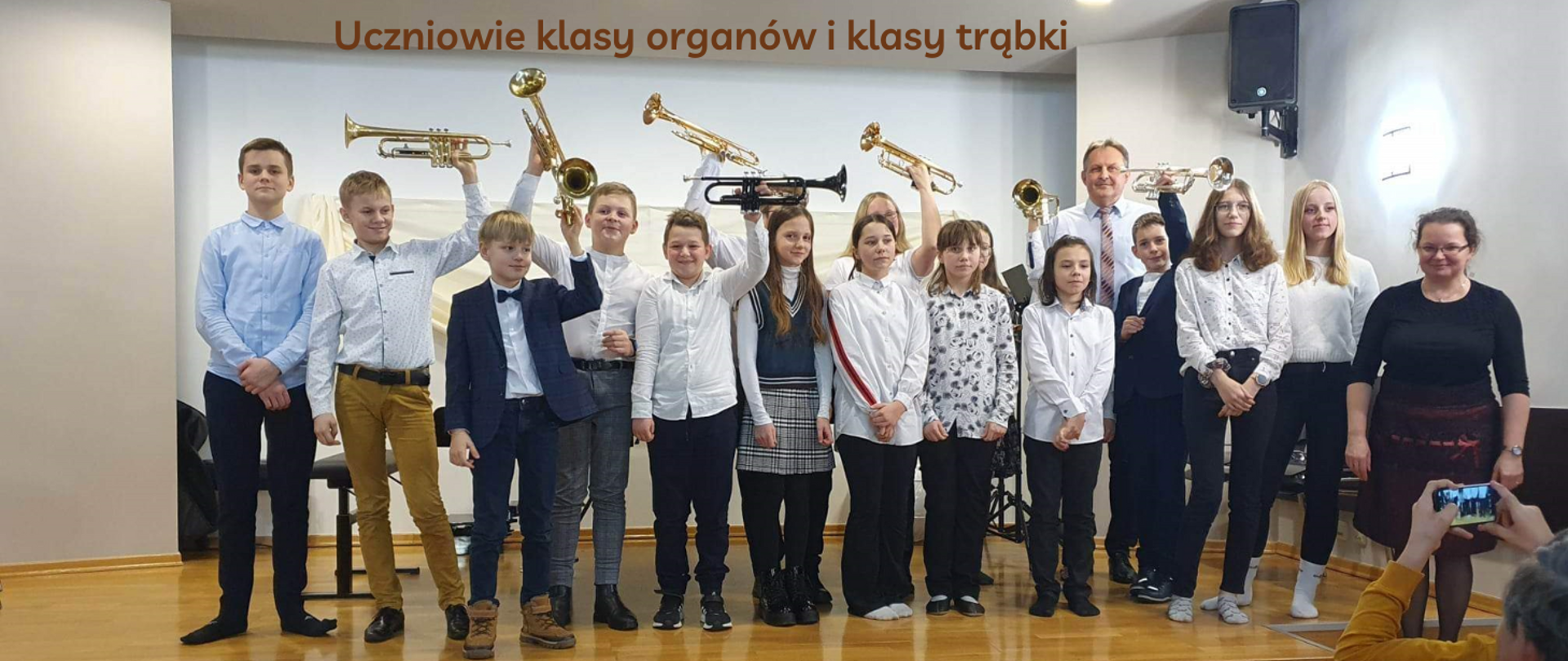 Na zdjęciu na scenie grupa uczniów. Wszyscy w odświętnych strojach galowych. Znajdziemy tutaj zarówno chłopców, jak i dziewczynki. Część z nich posiada trąbkę, którą unieśli do góry ponad głowę.