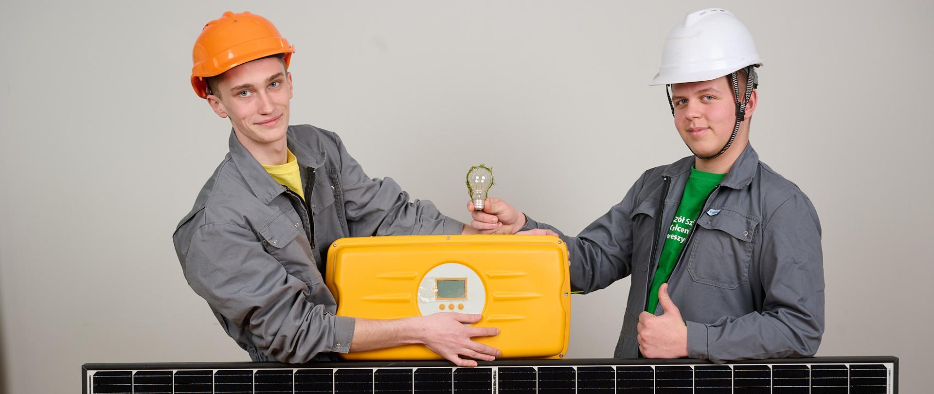Dwóch chłopców wspartych na panelu fotowoltaicznym. W ręku jeden trzyma żarówkę, a drugi podpiera walizkę ze sprzętem. Na głowach mają kaski ochronne.