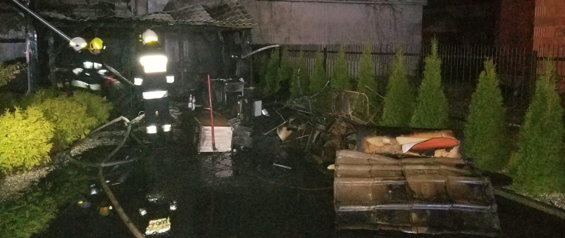 Zdjęcie przedstawia trawnik posesji na którym doszło do pożaru drewutni i dachu budynku mieszkalnego. Na trawie znajdują się spalone elementy wyposażenia znajdujące się w budynkach gospodarczych. Dodatkowo po lewej stornie na zdjęciu widać przystawioną do dachu drabinę oraz trzech strażaków. Strażacy ubrani w ubrania specjalne w kolorze ciemnym z elementami odblaskowymi. Na głowach mają białe i żółte hełmy. Zdjęcie wykonane w porze nocnej. 