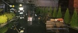 Zdjęcie przedstawia trawnik posesji na którym doszło do pożaru drewutni i dachu budynku mieszkalnego. Na trawie znajdują się spalone elementy wyposażenia znajdujące się w budynkach gospodarczych. Dodatkowo po lewej stornie na zdjęciu widać przystawioną do dachu drabinę oraz trzech strażaków. Strażacy ubrani w ubrania specjalne w kolorze ciemnym z elementami odblaskowymi. Na głowach mają białe i żółte hełmy. Zdjęcie wykonane w porze nocnej. 
