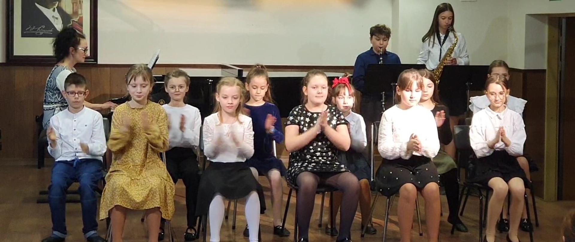W centrum klaszczący uczniowie ubrani na galowo siedzą na krzesełkach ustawionych na scenie, za nimi z lewej strony przy fortepianie siedzi kobieta w czarnych włosach w okularach, z prawej strony z tyłu stoi chłopiec grający na klarnecie, obok niego stoi dziewczyna grająca na saksofonie. Ściany koloru białego. Drewniana podłoga koloru brązowego, nad sceną widoczne 2 reflektory. Na tylnej ścianie wisi obraz z wizerunkiem Paderewskiego. Z prawej strony uchylone drzwi.