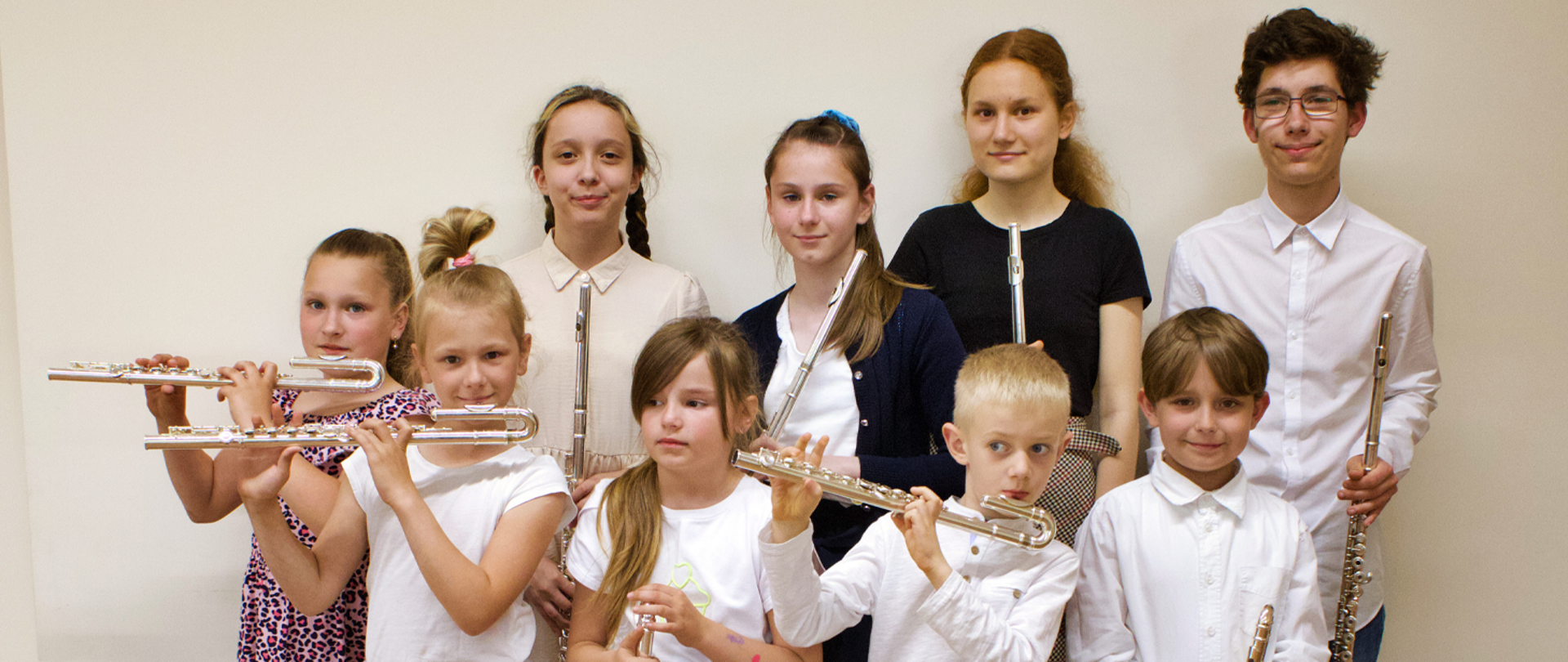 Grupa dzieci, siedem dziewczynek i dwóch chłopców stoją w dwóch rzędach na tle jasnej ściany. Wszyscy trzymają w rękach flety,troje z nich gra na nich w trakcie robienia zdjęcia.