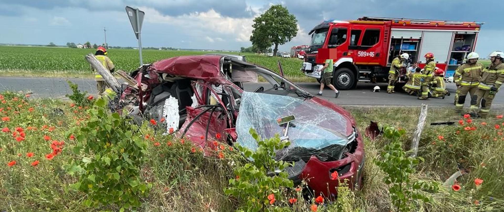 rozbity przód i bok samochodu stoi na poboczu drogi za nim na drodze samochód strażacki oraz strażacy