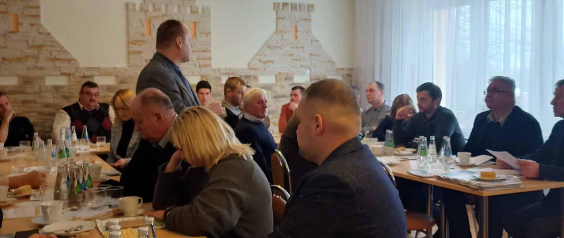 Sala konferencyjna wypełniona uczestnikami siedzącymi przy stołach. Mężczyzna w szarym garniturze stoi i przemawia do grupy. Na stołach widać filiżanki, wodę i egzemplarze biuletynu.