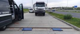 Na pierwszym planie (od lewej): prawy bok oznakowanego furgonu mazowieckiej Inspekcji Transportu Drogowego i inspekcyjne wagi umieszczone w zagłębieniu nawierzchni zatoki kontrolnej. W tle: zatrzymana do kontroli wywrotka stoi przed rozłożonymi wagami. Jej kierowca przygotowuje się do przejazdu przez urządzenia służące do pomiaru rzeczywistych nacisków osi pojazdu na drogę.