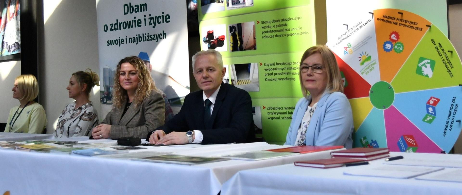 Mężczyzna siedzi pomiędzy dwoma kobietami za stołem stanowiska KRUS, za nimi rollupy KRUS i Koło Bezpieczeństwa - gra prewencyjna do losowania pytań i nagród.