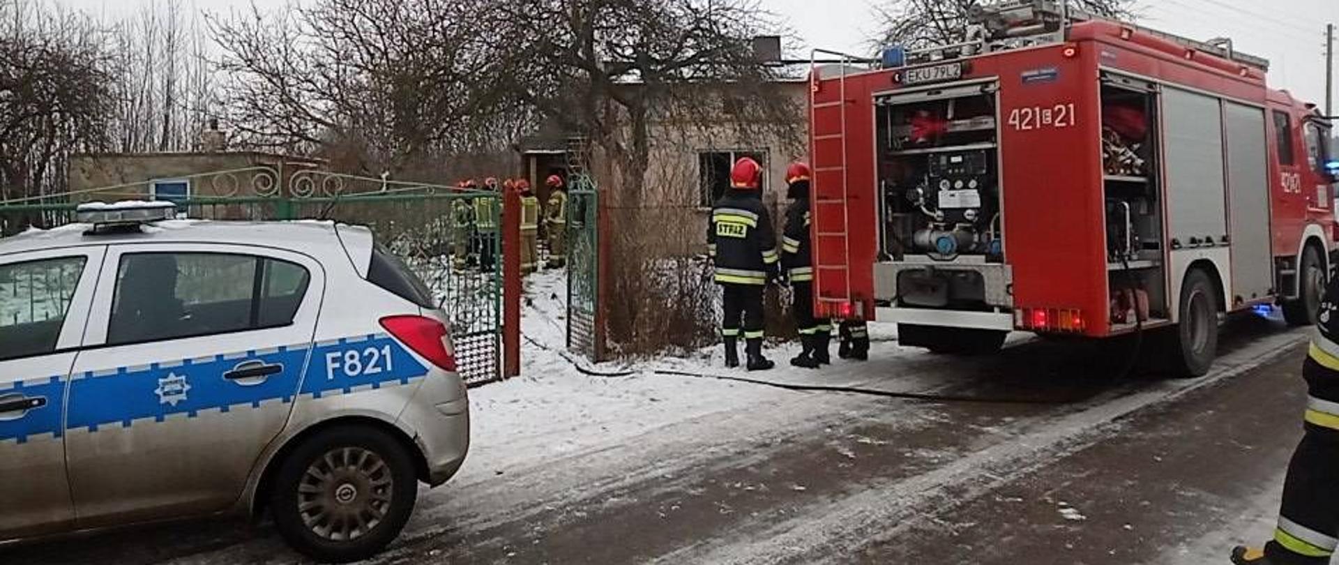 pożar budynku mieszkalnego. Przed budynkiem 5 strażaków ubranych w ubranie specjalne w kolorze żółtym i czerwone hełmy. Przed palącym się budynkiem stoi czerwony pojazd specjalny straży pożarnej oraz radiowóz policyjny. Przed pojazdem gaśniczym straży pożarnej znajdują się strażacy prowadzący działania ratowniczo - gaśnicze . Do budynku objętego po9żarem ciągnie się rozłożona linia gaśnicza. 