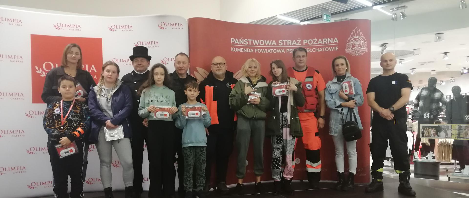 na zdjęciu widzimy laureatów konkursu czad i ogeiń obudź czujność wraz z przedstawicielami pogotowia, kominiarzem oraz prowadzącymi akcję w tle logo straży oraz galerii olimpia