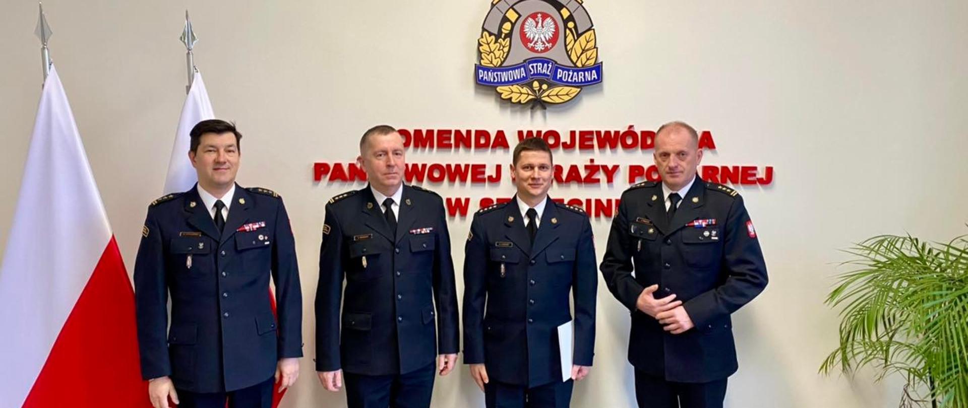 Na zdjęciu 4 funkcjonariuszy, od lewej: zastępca zachodniopomorskiego Komendanta Wojewódzkiego PSP st. bryg. Marek Popławski, Komendant Powiatowy PSP w Kołobrzegu st. bryg. Krzysztof Makowski, p.o. Zastępcy Komendanta Powiatowego w Kołobrzegu st.kpt. Krzysztof Azierski, Zachodniopomorski Komendant Wojewódzki PSP w Szczecinie st. bryg. Jarosław Tomczyk
W tle reprezentacyjna ścianka sali narad z napisem "Komenda Wojewódzka Państwowej Straży Pożarnej w Szczecinie", po lewej stronie flagi Polski, po prawej stronie kwiat.