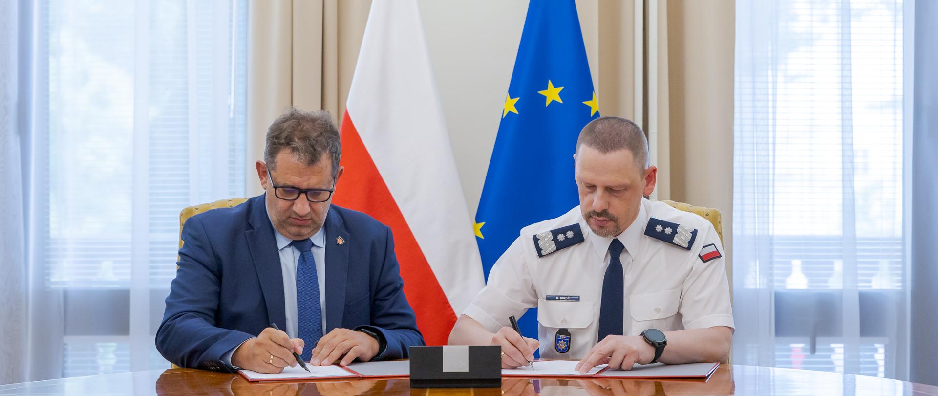 Dwie osoby siedzą przy stole i podpisują dokumenty, a w tle widoczne są flagi Polski i Unii Europejskiej.