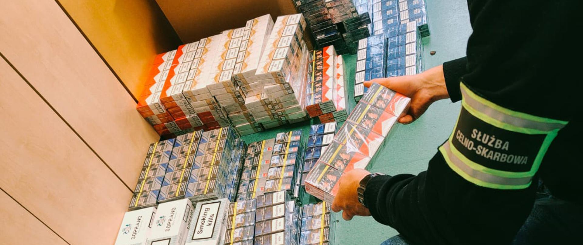 Pokój biurowy, przy szafie na podłodze poukładane zafoliowane opakowania papierosów z różnymi etykietami, przed nimi kuca funkcjonariusz Służby Celno-Skarbowej, jedno z opakowań trzyma w dłoniach
