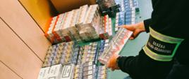 Pokój biurowy, przy szafie na podłodze poukładane zafoliowane opakowania papierosów z różnymi etykietami, przed nimi kuca funkcjonariusz Służby Celno-Skarbowej, jedno z opakowań trzyma w dłoniach