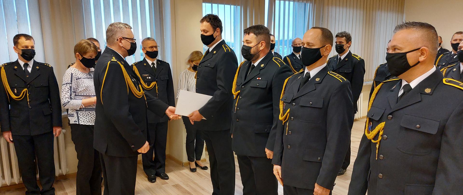 Na zdjęciu widoczni są funkcjonariusze KM PSP w Tarnowie u umundurowaniu galowym podczas uroczystej zbiórki. Komendant Miejski PSP w Tarnowie wręcza strażakom awanse. Zbiórka ma miejsce w Sali Narad KM PSP w Tarnowie 
