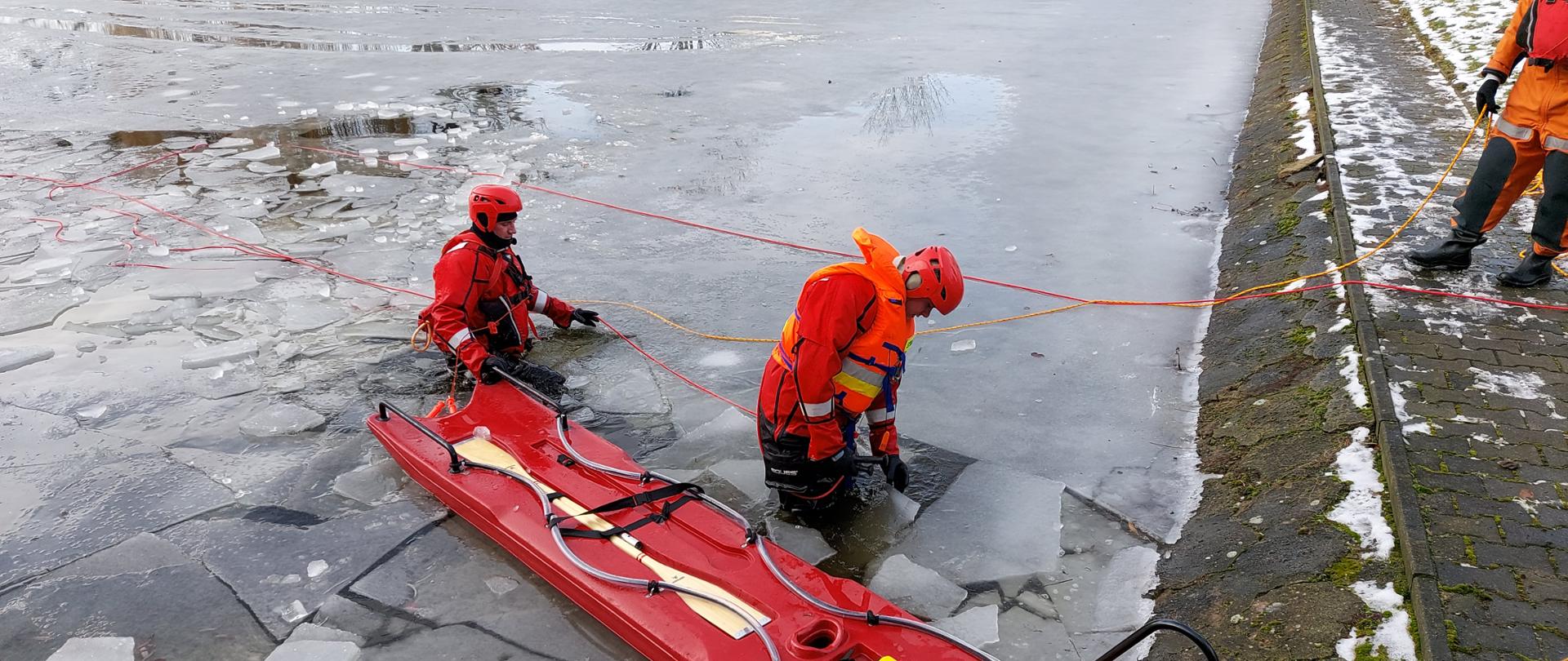 Dwóch strażaków stojących w zamarzniętym zbiorniku wodnym.