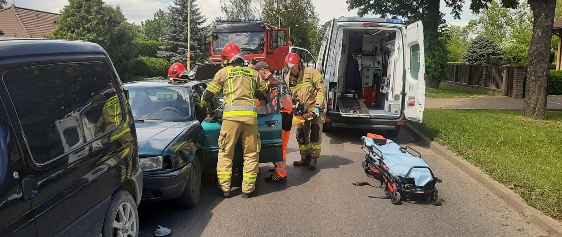Strażacy z ratownikami medycznymi ewakuują kierowcą z pojazdu, po prawej tył karetki z otwartymi drzwiami 