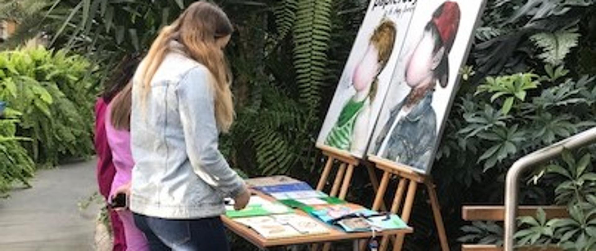 Osoby odwiedzające wystawę prac finalistów etapu wojewódzkiego konkursu pt.: „Palić, nie palić – oto jest pytanie?”, na stole leżą lapbooki wykonane przez uczniów klas V, uczestników konkursu. Wystawa odbywa się w Ogrodzie Zimowym Wielkopolskiego Centrum Onkologii.