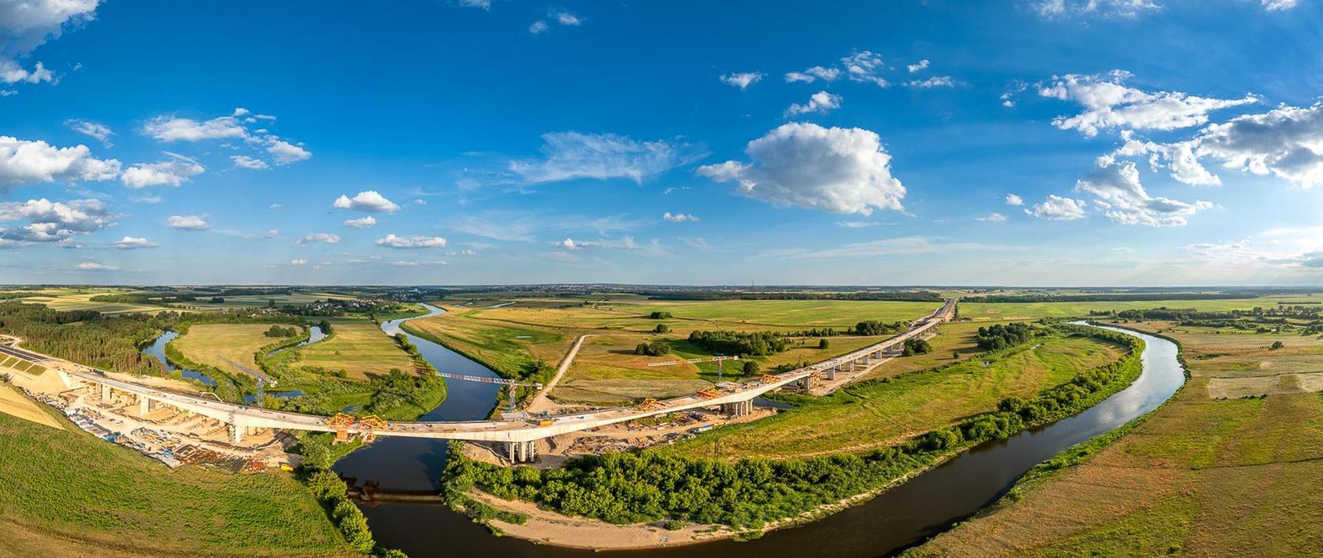Widok z lotu ptaka na wielką plac budowy mostu nad meandrującą rzeką. Rzeka wygina się przez krajobraz, zielone pola i skupiska drzew po obu stronach. Na placu budowy widać wiele żurawi, kilka częściowo ukończonych odcinków mostu z widocznymi rusztowaniami oraz liczne pojazdy budowlane. Niebo jest bezchmurne, co sugeruje dobre warunki pogodowe do prac budowlanych.