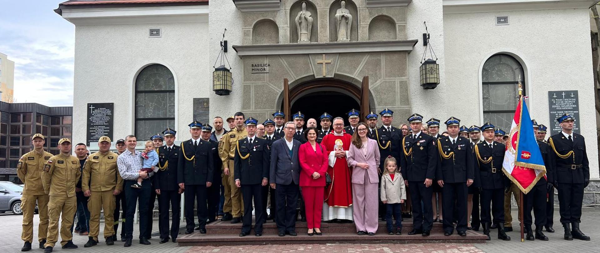 Zbiórka strażaków oraz osób uczestniczących w mszy świętej z okazji dnia strażaka. Strażacy w mundurach wyjściowych oraz służbowych. Stoją przed kościołem.