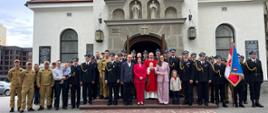 Zbiórka strażaków oraz osób uczestniczących w mszy świętej z okazji dnia strażaka. Strażacy w mundurach wyjściowych oraz służbowych. Stoją przed kościołem.