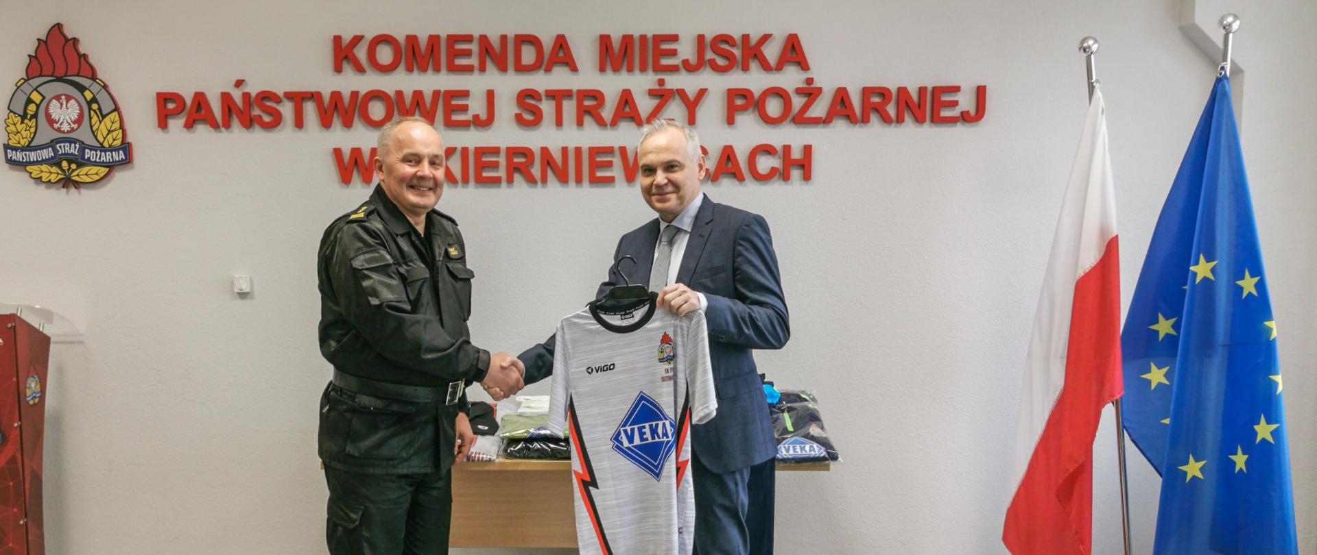 Na zdjęciu moment przekazania stroju sportowego przez mężczyznę w garniturze, który podał dłoń strażakowi w mundurze w kolorze czarnym. Przekazanie odbywa się na tle czerwonego napisu Komenda Miejska Państwowej Straży Pożarnej w Skierniewicach, loga psp, flag polski i unii europejskiej.