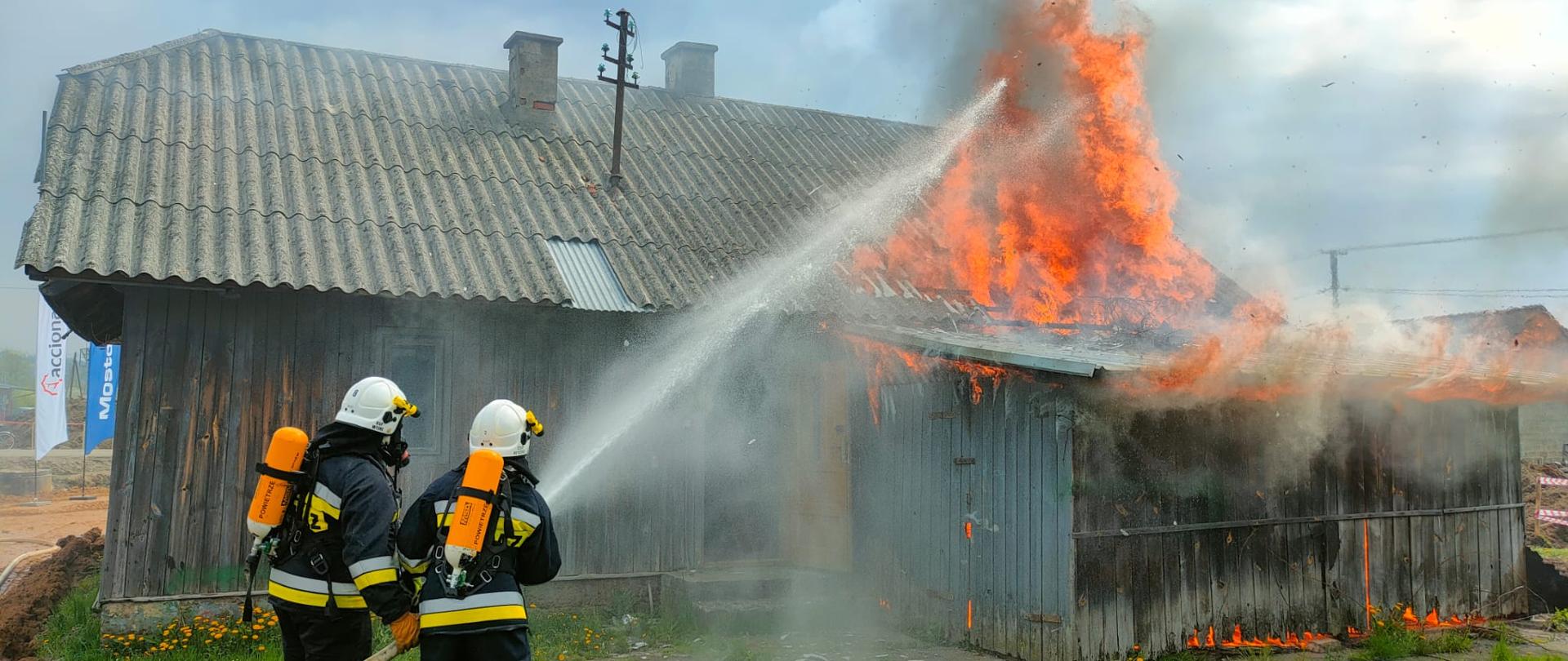 Na zdjęciu widoczny budynek drewniany objęty pożarem. Przed budynkiem dwóch strażaków w ubraniach ochronnych oraz aparatach OUO z linia gaśniczą - gaszą budynek.