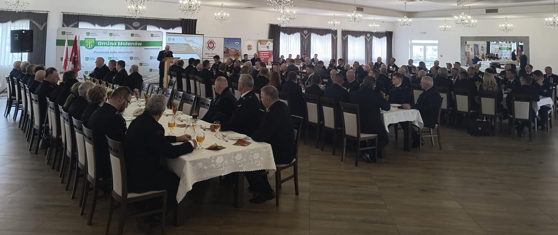 Uczestnicy odprawy szkoleniowej siedzą na krzesłach przy stołach słuchając przemawiającego druha Grzegorza Ciesielskiego. 