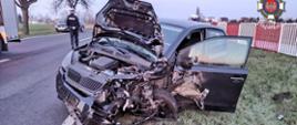 Zdjęcie przedstawia zniszczony samochód osobowy stojący częściowo na jezdni, a częściowo na trawie. Po lewej stronie pojazdu tyłem stoi policjant. Z prawej strony biało- czerwone barierki ochronne